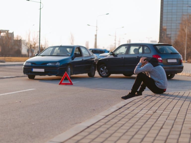 A man sits on the curb with his head down next to two collided cars in an accident.