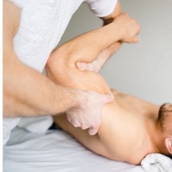 A male laying on a therapy table having his left arm worked on by a therapist.