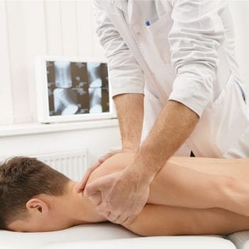A photo of a male laying on a theapy table having his left shoulder stretched by a provider.