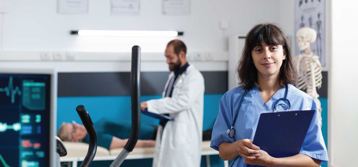 A medical provider asses a patient in the background while a medical assistant stands in front and looks forward while holding a chart.